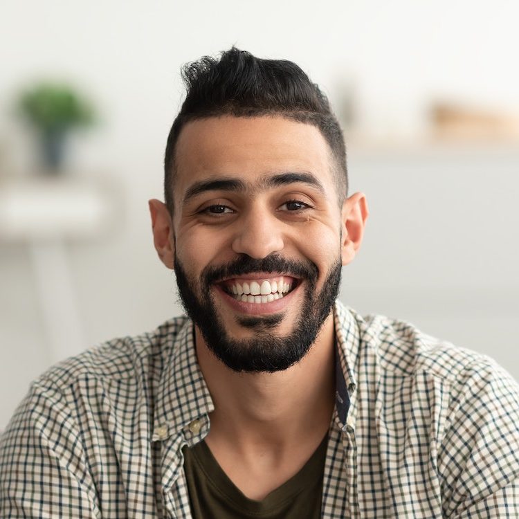 Portrait of handsome young arab man smiling and looking at camera sitting on sofa at home e1682562032200.jpg